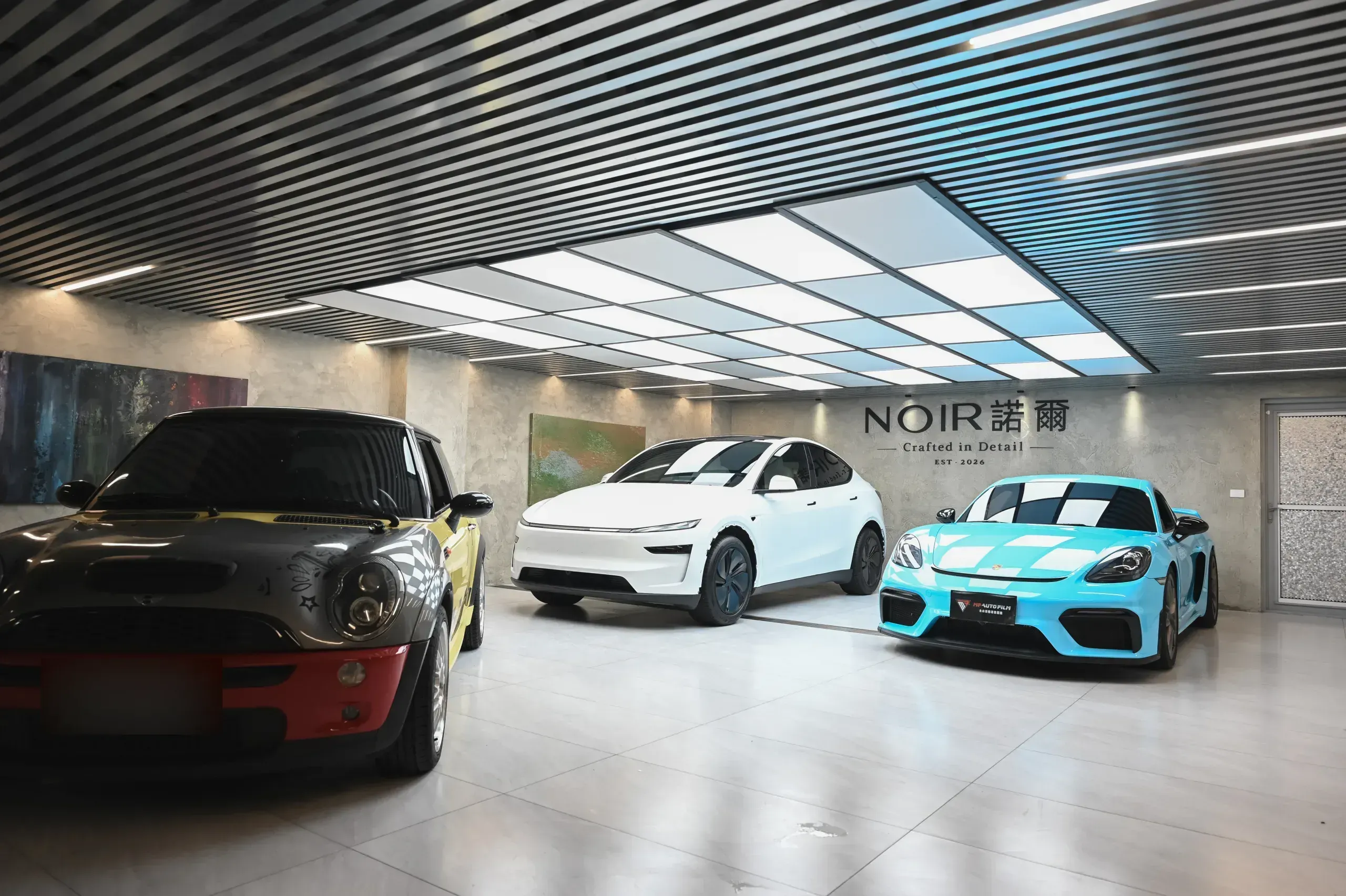NOIR 諾爾施工棚 — MINI Clubman、Tesla Model Y、Porsche Cayman 於光柵天花下，背景為 NOIR 諾爾雕刻牆